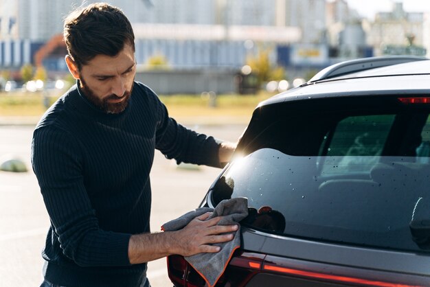 Cintura para cima vista de retrato de um homem concentrado e satisfeito limpando a parte de trás de seu carro novo, enquanto passava um tempo na rua durante o dia ensolarado de verão. Foto