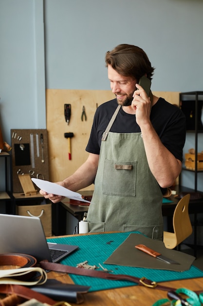 Cintura vertical para arriba retrato del artesano masculino sonriente que usa la computadora portátil y que habla por el teléfono inteligente mientras trabaja ...