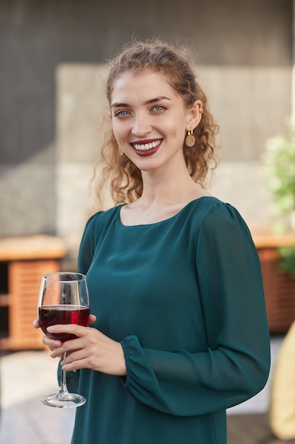 Cintura vertical para arriba retrato de mujer joven sonriente sosteniendo una copa de vino y mientras disfruta de la fiesta al aire libre