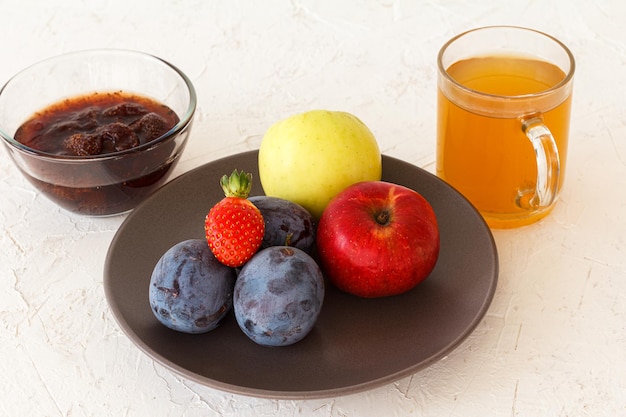 Ciruelas maduras recogidas manzanas una fresa en el plato y un tazón de vidrio con mermelada y una taza de té