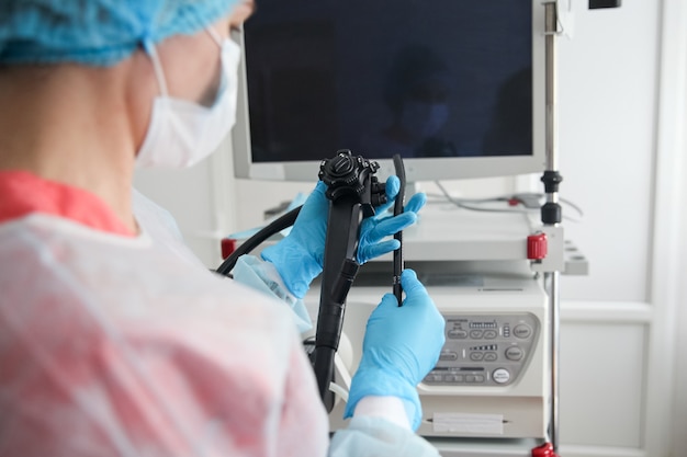 Una cirujana endoscopista con traje de protección, gorra, máscara y guantes sostiene un endoscopio frente a un monitor