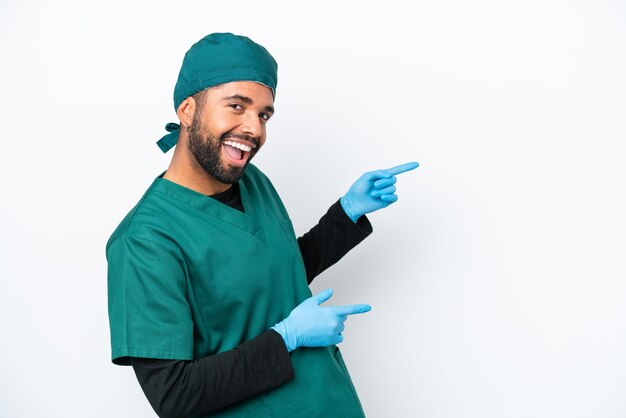 Cirujano brasileño con uniforme verde aislado de fondo blanco señalando con el dedo al costado y presentando un producto