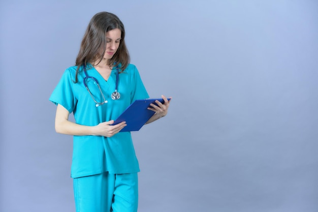 Cirujano femenino en exfoliantes de color aguamarina mirando el portapapeles sobre fondo azul.