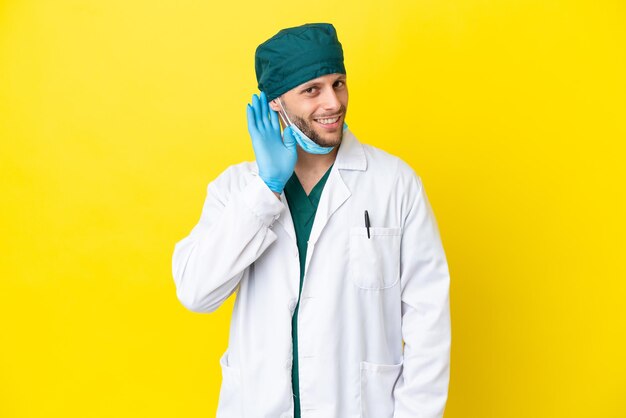 Cirujano hombre rubio en uniforme verde aislado sobre fondo amarillo escuchando algo poniendo la mano en la oreja