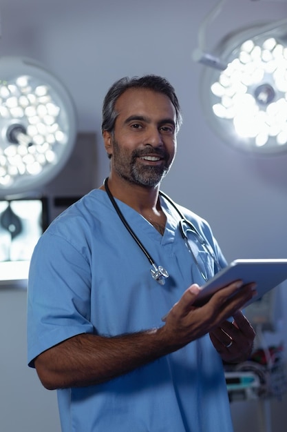 Foto cirujano masculino maduro mirando a la cámara mientras usa una tableta digital en la sala de operaciones del hospital