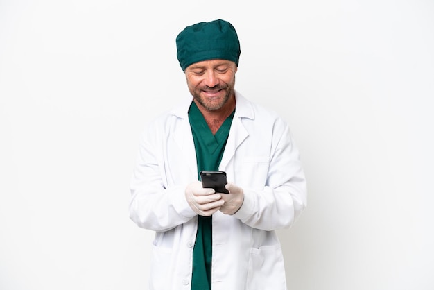 Cirujano de mediana edad con uniforme verde aislado de fondo blanco enviando un mensaje con el móvil