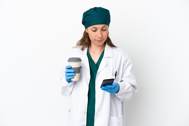 Cirujano mujer caucásica en uniforme verde aislado sobre fondo blanco sosteniendo café para llevar y un móvil
