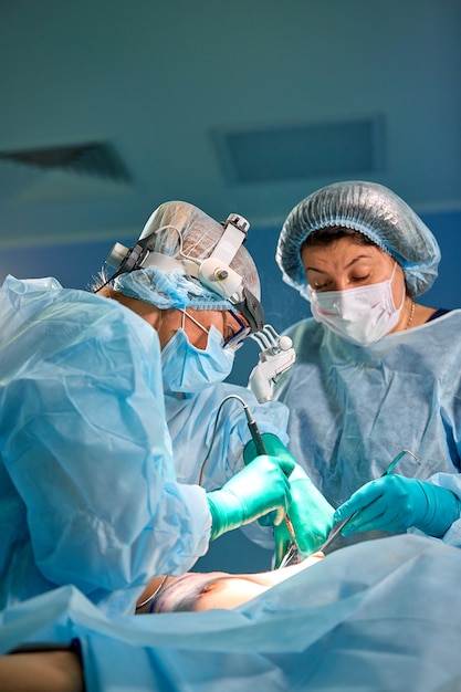 Cirujano que realiza cirugía estética en el quirófano del hospital. Cirujano en máscara con lupas durante el procedimiento médico. Aumento de senos