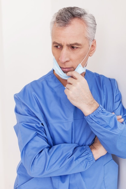 Cirujano reflexivo. Médico senior de cabello gris deprimido quitándose la mascarilla quirúrgica mientras se inclina contra la pared