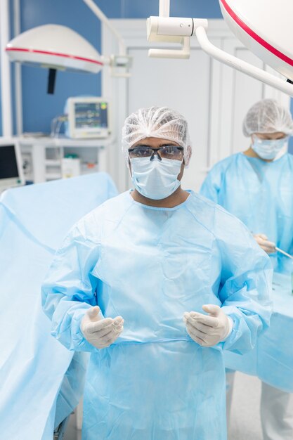 Cirujano en uniforme protector, máscara, guantes y anteojos mirándolo contra su asistente preparando instrumentos médicos antes de la operación