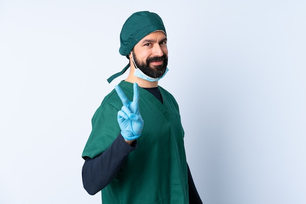 Cirurgião homem de uniforme verde sobre parede isolada, sorrindo e mostrando sinal de vitória