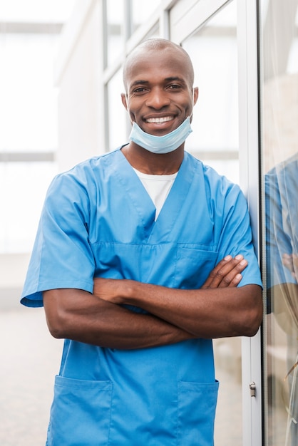 Cirurgião jovem e bem sucedido. Jovem médico africano confiante de uniforme azul olhando para a câmera e sorrindo enquanto mantém os braços cruzados