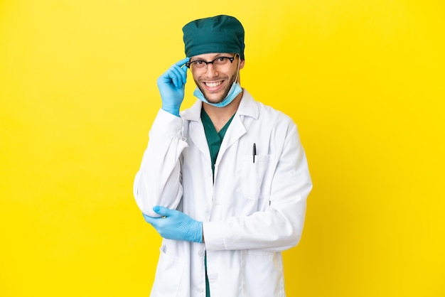 Cirurgião loiro de uniforme verde isolado em fundo amarelo com óculos e feliz