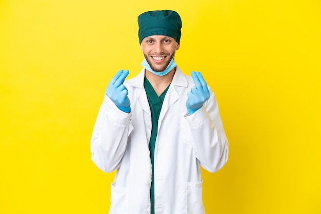 Cirurgião loiro de uniforme verde isolado em fundo amarelo fazendo gesto de dinheiro