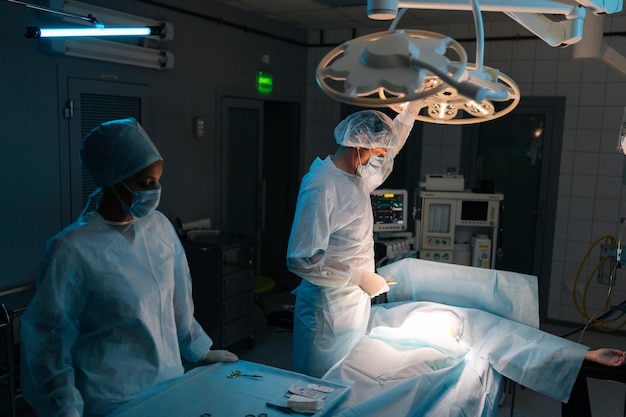 Cirurgião masculino em uniformes cirúrgicos e máscaras ajustando a luz antes da operação no escuro operando