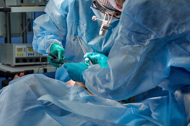 Cirurgião realizando cirurgia estética na sala de cirurgia do hospital Cirurgião em máscara usando lupas durante procedimento médico Aprimoramento de aumento de mama Filtro azul de fundo azul