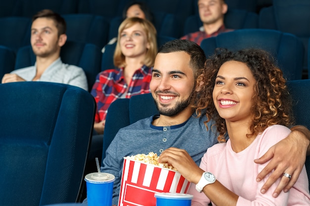 ¡Cita nocturna en el cine! Joven pareja multicultural feliz comiendo palomitas y viendo películas juntos en el cine