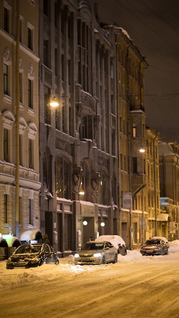 Ciudad de invierno por la noche calle de la ciudad vacía con coche bajo la nieve después de las nevadas y luces de la calle brillante