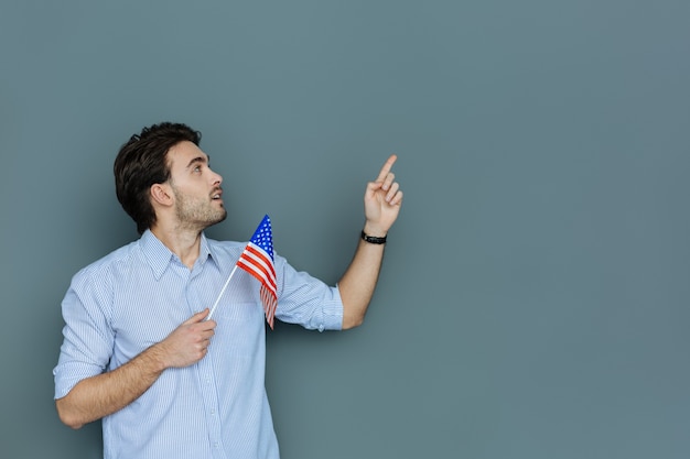 Ciudadano americano. Agradable agradable hombre feliz sosteniendo la bandera estadounidense y mirando en la dirección de su mano mientras es un patriota estadounidense