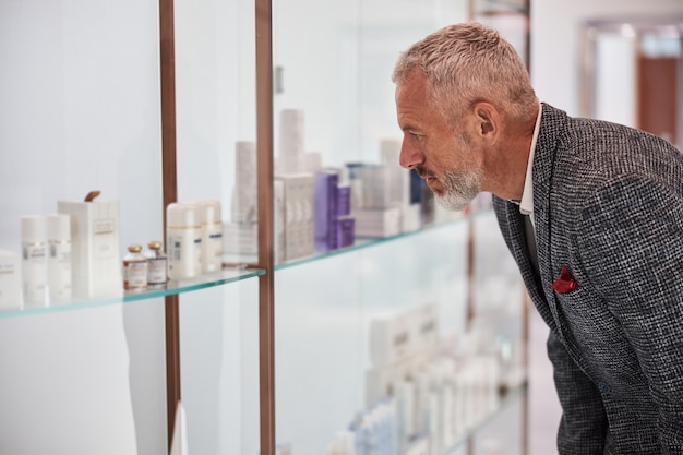 Ciudadano anciano serio eligiendo productos cosméticos de un estante en una tienda bien iluminada