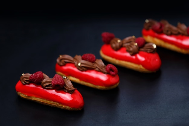 Éclair de bolos de creme com cobertura de chocolate colorido e framboesas em um fundo preto