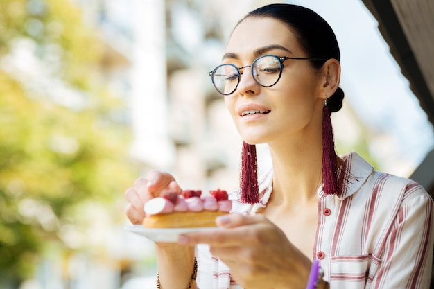 Éclairs amorosos. Mulher alegre e relaxada apreciando a deliciosa sobremesa de framboesa e sorrindo enquanto olha gentilmente para ela