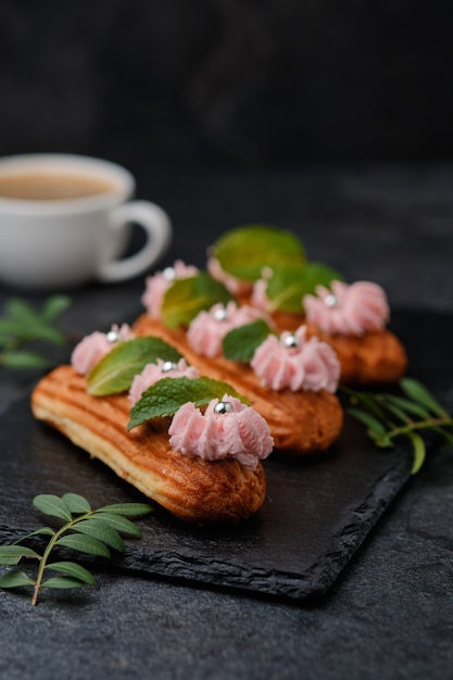 Éclairs com creme rosa, decorados com folhas de hortelã. Sobremesa em um prato de ardósia preta. Bolos e café expresso em um fundo escuro.