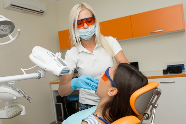 Clareamento dos dentes pelo dispositivo de clareamento UV dental, assistente dental cuidando do paciente, olhos protegidos com óculos. Tratamento clareador com luz, laser, flúor. Clareamento artificial dos dentes