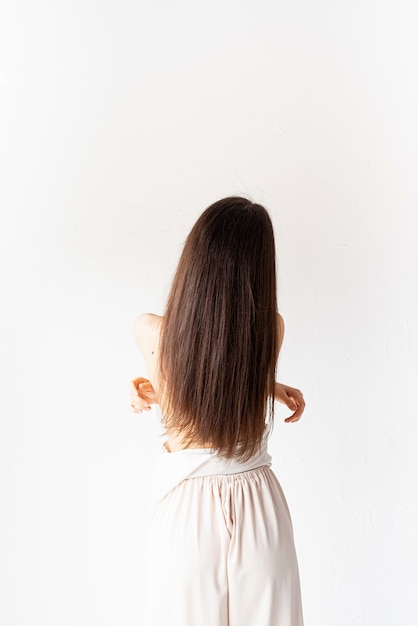 Claro e arejado. mulher jovem e bonita com cabelo comprido em roupas brancas aconchegantes em fundo branco, vista de trás