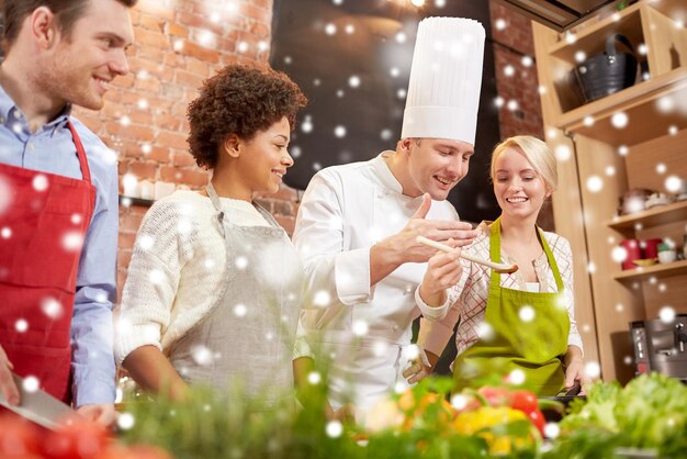 Foto clase de cocina, concepto culinario, de comida y de personas - feliz grupo de amigos y chef masculino cocinando en la cocina sobre el efecto de la nieve