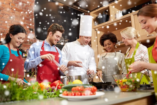 Foto clase de cocina, concepto culinario, de comida y de personas - feliz grupo de amigos y chef masculino cocinando en la cocina sobre el efecto de la nieve