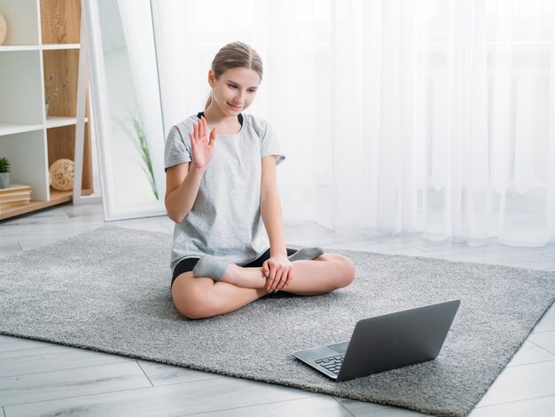 Clase de yoga en línea Entrenamiento en el hogar Fitness para principiantes Blog de videos deportivos Chica relajada saludando agitando la mano en la computadora portátil sentada con las piernas cruzadas en el suelo en un interior claro