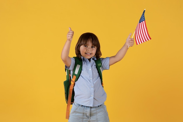 Clases preescolares internacionales Lindo niño pequeño con mochila sosteniendo la bandera de EE. UU. Y mostrando el pulgar hacia arriba gesto espacio libre