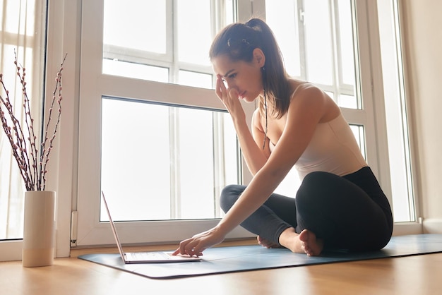 Clases de yoga en línea Chica de yoga positiva haciendo práctica matutina frente a la computadora portátil en casa