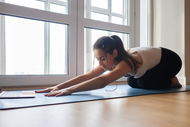 Clases de yoga en línea Chica de yoga positiva haciendo práctica matutina frente a la computadora portátil en casa