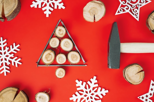 Clavos largos y cortes de madera o losas de ramas, dispuestos en forma de árbol de Navidad con martillo y copos de nieve de madera sobre fondo rojo. Endecha plana