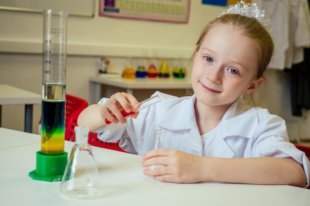 Clevere blonde Schulchemikerin in Brille und weißem medizinischem Kittel, die wissenschaftliche Experimente Chemie macht und verschiedene chemische Lösungen im Laborklassenzimmer mischt