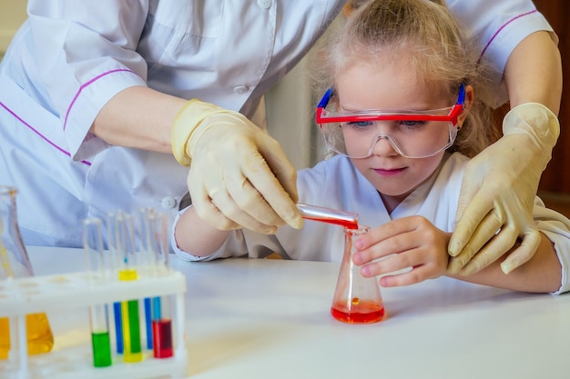 Clevere blonde Schulmädchen-Chemikerin mit Brille und weißem Arztkittel, die wissenschaftliche Experimente in der Chemie durchführt und verschiedene chemische Lösungen im Laborklassenzimmer mischt