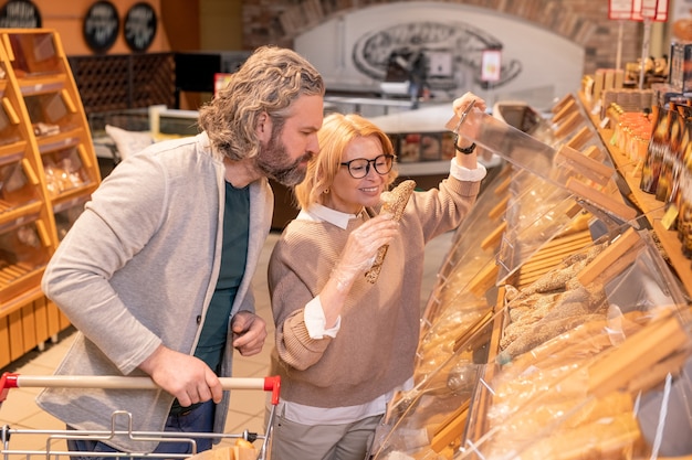 Clienta feliz tomando pan fresco de la pantalla de madera mientras compra productos alimenticios con su esposo en el supermercado
