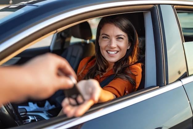 Una clienta optimista sentada en un auto nuevo y tomando las llaves de un distribuidor de cultivos en un concesionario, una mujer hace compras en la sala de exposición de autos, se sienta dentro de un hermoso auto y mira al agente de ventas