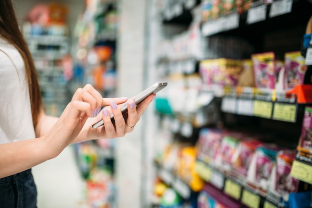 Clienta con teléfono en mano, tienda de alimentos. Mujer usa smartphone en supermercado