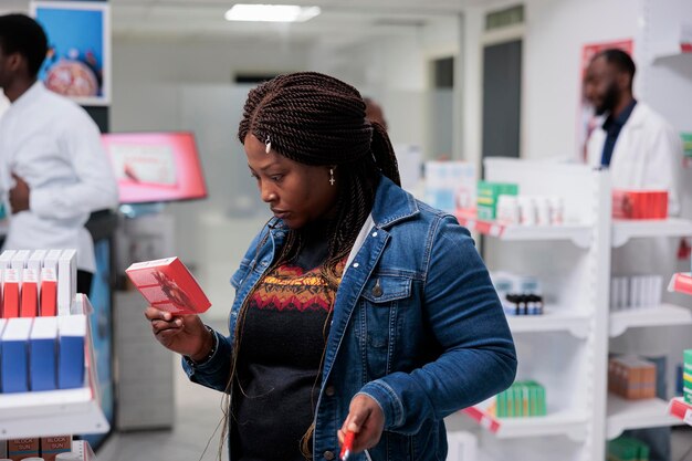 Cliente afro-americano comprando medicamentos para o coração na farmácia, lendo instruções, segurando o pacote de comprimidos, parado no corredor da farmácia. cliente escolhendo vitaminas, tiro médio