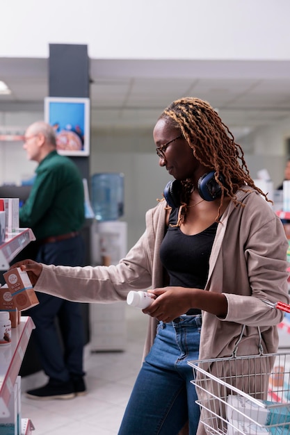 Cliente afro-americano segurando cesta de compras olhando para prateleiras cheias de produtos de saúde, comprando creme spf. Cliente optando por comprar suplementos, vitamina para sistema imunológico. conceito de medicina