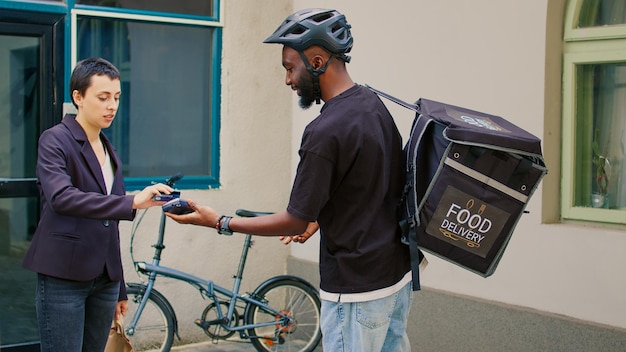 Cliente caucasiano pagando pedido de entrega de fastfood com cartão de crédito, pagamento sem contato pos na porta da frente do escritório. Correio de restaurante com mochila segurando terminal, dando refeição em saco de papel.