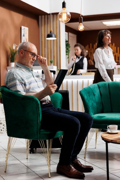 Un cliente caucásico anciano en el salón del hotel sentado en un cómodo sofá con una tableta digital Un anciano jubilado navegando por la red para actividades de vacaciones divertidas en su dispositivo inteligente en un agradable complejo turístico