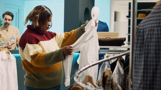 Cliente da loja de varejo examinando roupas em prateleiras, procurando comprar uma nova coleção da moda para aumentar o guarda-roupa. Mulher nova que verifica a mercadoria da loja de roupa no shopping moderno.