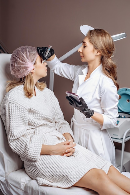 Cliente de boné e roupão recebe marcas no rosto com caneta nas mãos de cosmetologista experiente para fornecer serviços de cosmetologia