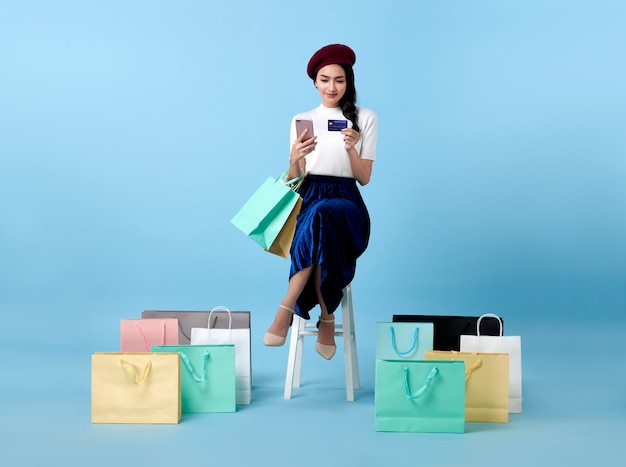 Cliente de linda mulher asiática sentado e carregando sacolas de compras com o uso de cartão de crédito e telefone celular nas mãos no espaço azul.