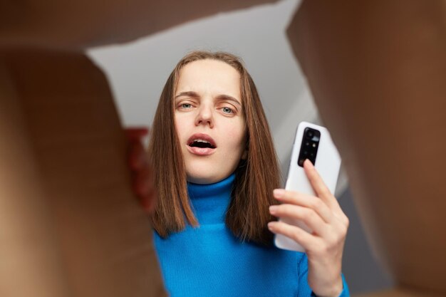 Cliente de mulher de cabelos castanhos assustada chocada vestindo gola alta azul desempacotando pacote olhando dentro do pacote com choque segurando a vista do telefone móvel do fundo da caixa