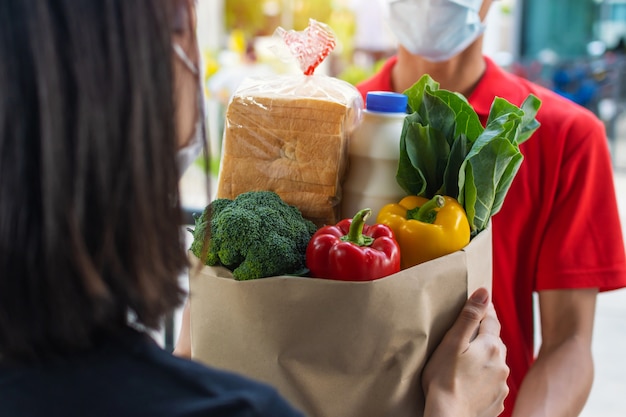 Foto cliente de mulher recebendo saco conjunto de alimentos frescos do homem de serviço de entrega de alimentos com máscara facial de proteção em uniforme vermelho na porta de casa, entrega expressa, quarentena, surto de vírus, conceito de entrega de alimentos